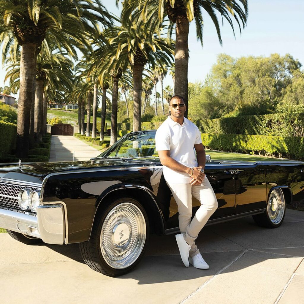 Russell Wilson modeling Good Man Brand Men's clothing in front of a Black lincoln Town Car